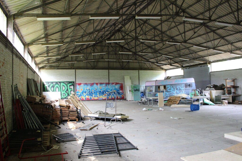 a photograph of the interior of an abandoned warehouse in belgium. there is a caravan, emergency beds and other stuff on the ground.