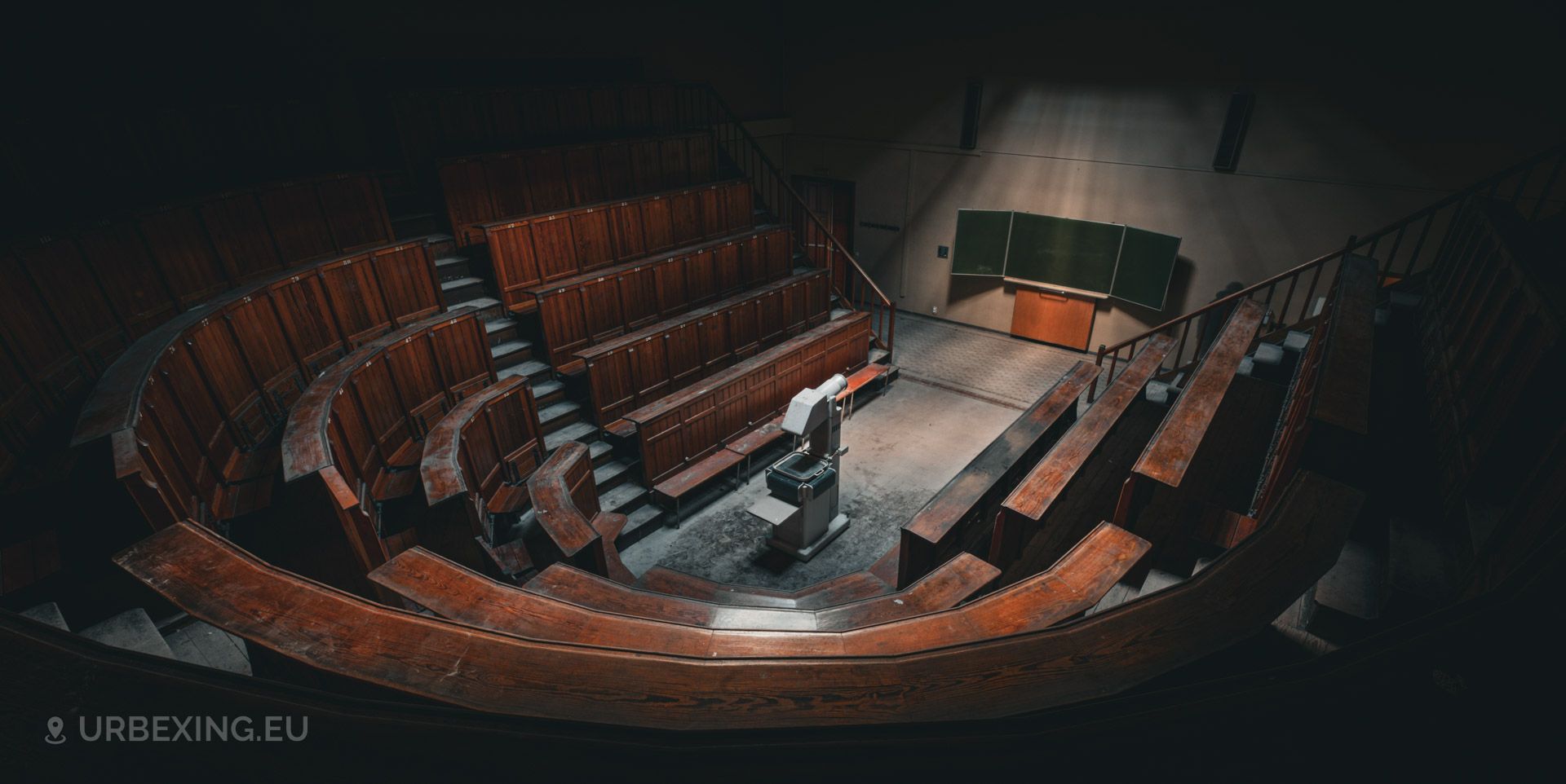 a photograph taken in an abandoned medical university. the photograph shows the overview of an auditorium, rows of wooden benches, a chalk board and an old projector