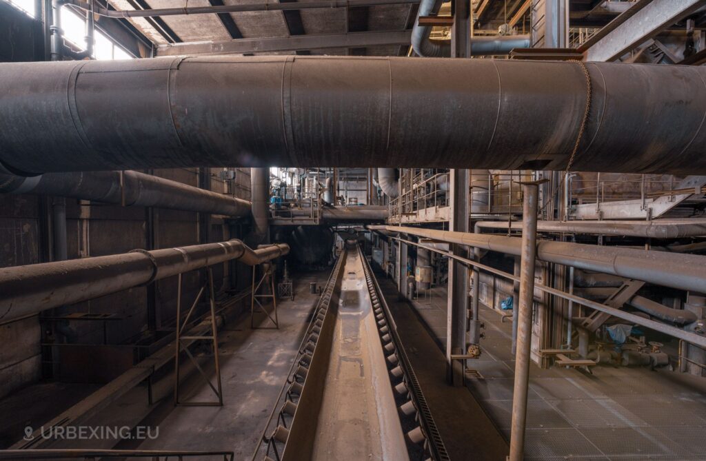 A picture inside the boiler house of an abandoned power plant. The picture show a conveyor belt, tubes and other machinery.