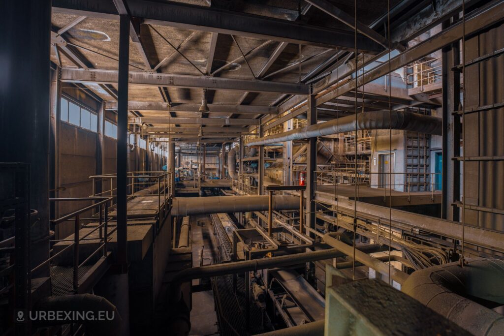 A picture inside of the boiler house at an abandoned power plant. The picture shows a conveyor belt, tubes, fire extinguisher, an elevator and other machinery.