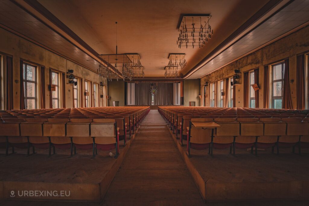 a photograph taken inside an abandoned police academy. the photo is taken in the auditorium and shows many rows of wooden folding chairs, several chandeliers and a large logo of the german bundehspolizei
