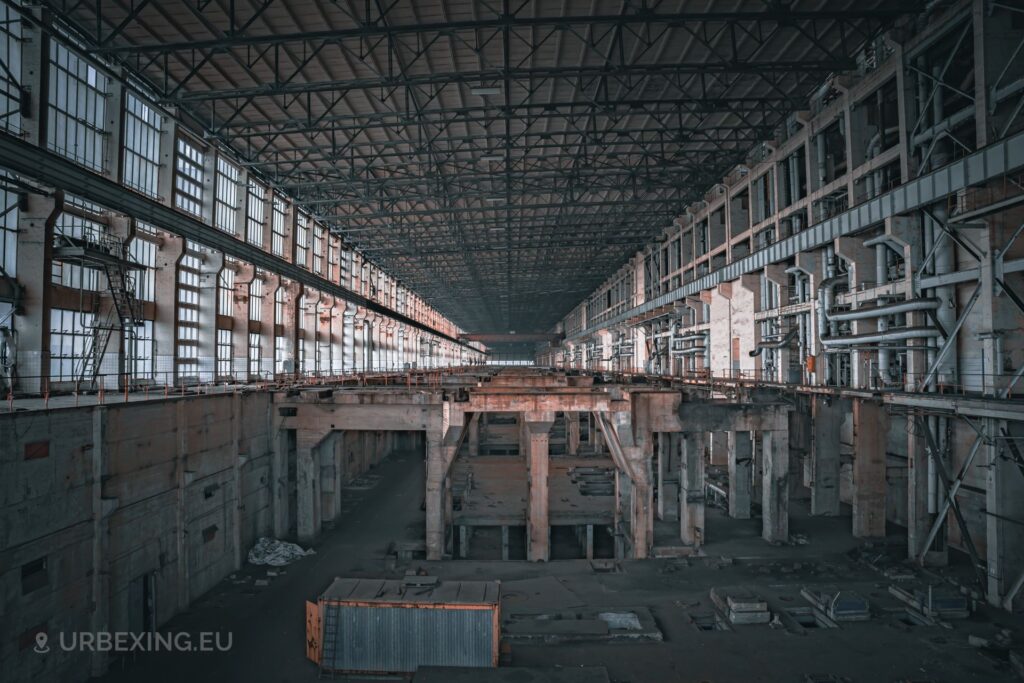 a photo of a turbine hall at a former power plant in germany. the image shows an empty hall with a crane in the distance and a yellow shipping container.