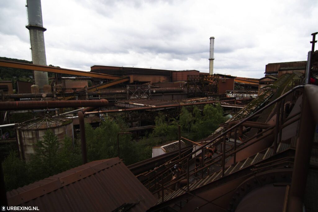 een foto van de verlaten staalfabriek in Liege, Belgie. Op de foto zijn meerdere buizen, een grote hal en schoorstenen te zien.
