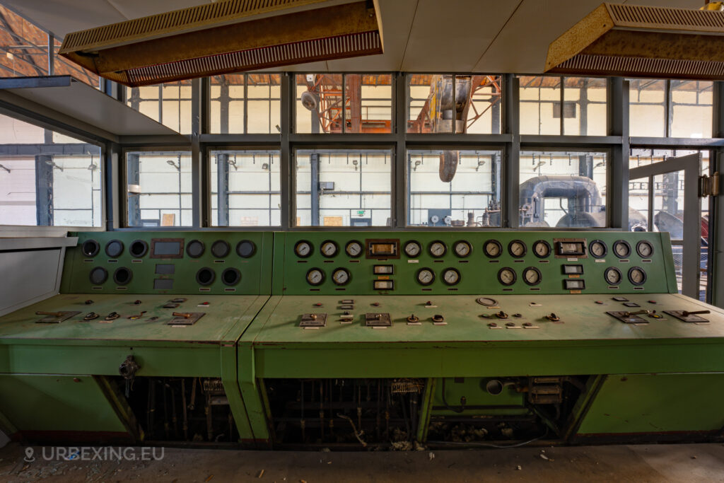 a photograph of a green control panel in a former power plant. a turbine can be seen in the background.