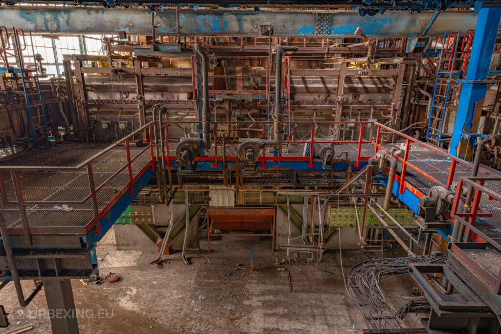 a photograph taken inside an abandoned glass factory called "de glasfabriek schiedam". The photograph shows a green control panel, a glass oven and several blowers used to heat the oven.