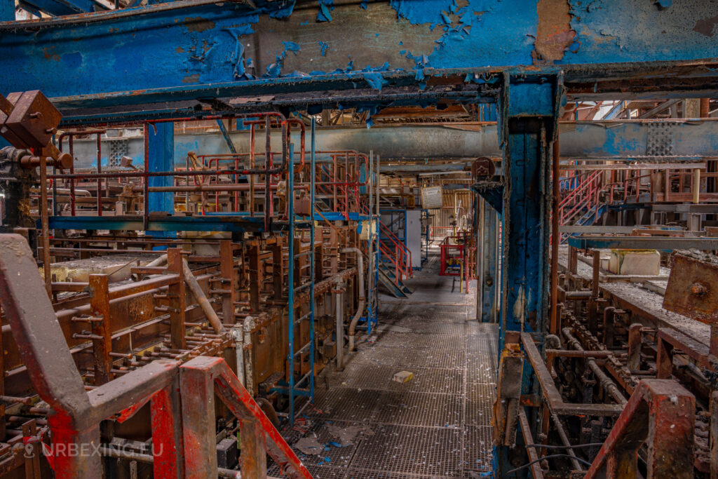 a photograph taken inside an abandoned glass factory called "de glasfabriek schiedam". The photograph shows an overview of the ovens inside the glass factory, there are several machines colored in red and blue