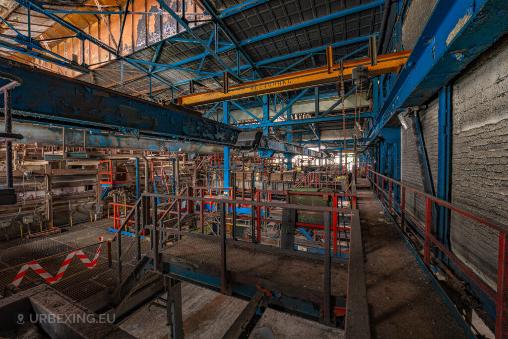 a photograph taken inside an abandoned glass factory called "de glasfabriek schiedam". The photo shows hall with a yellow crane hanging from the ceiling, multiple catwalks colored in red and blue