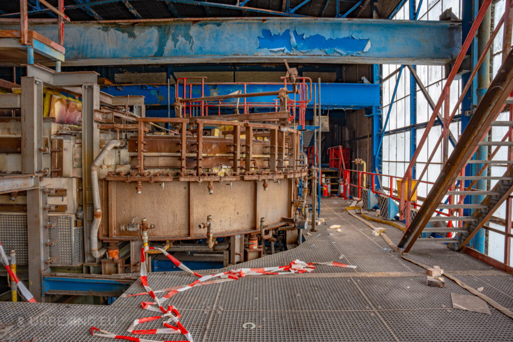a photograph taken inside an abandoned glass factory called "de glasfabriek schiedam". The photograph shows a transporting system for hot glass bottles. The factory is painted in red and blue coloring