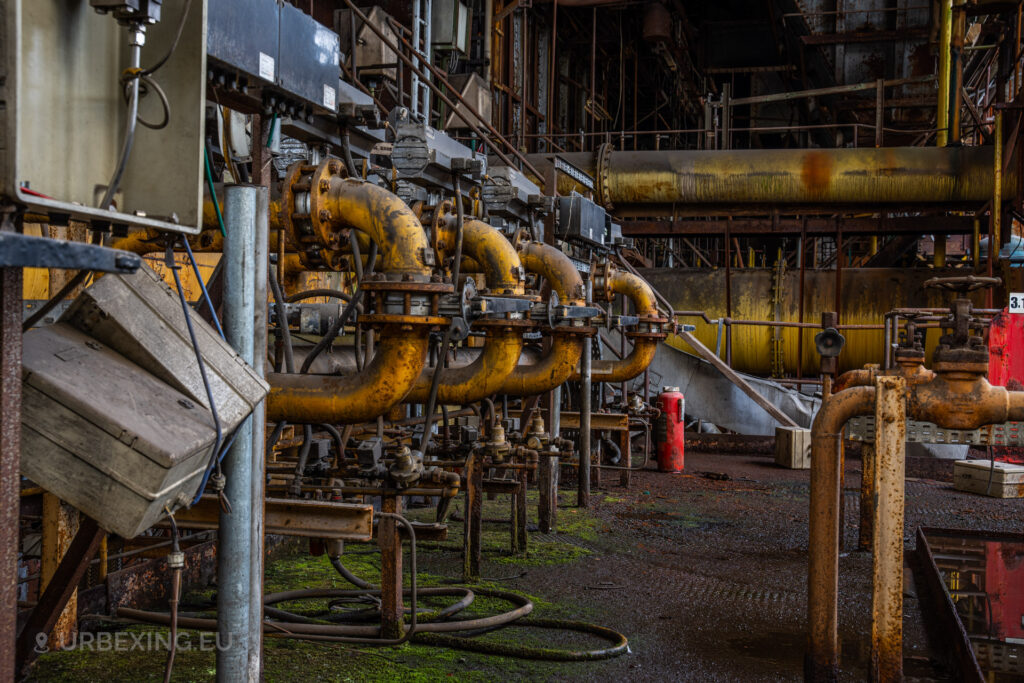 a photograph taken in at an urbex location called blue power plant. the photograph shows several yellow gas pipes. it is decaying, rust can be seen everywhere