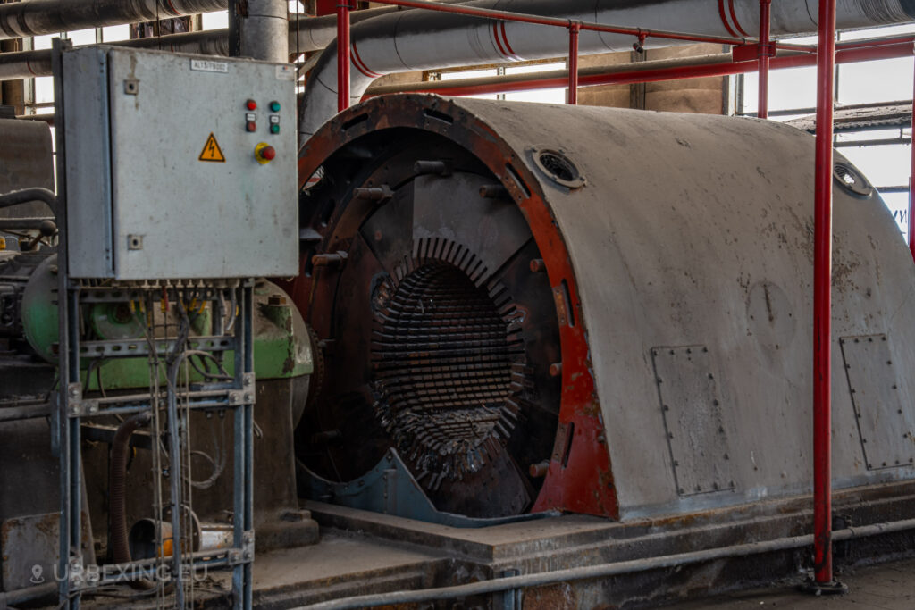 a photograph taken in at an urbex location called blue power plant. the photograph shows a close up of one of the red alternators in the turbine hall. the alternator has been broken open.