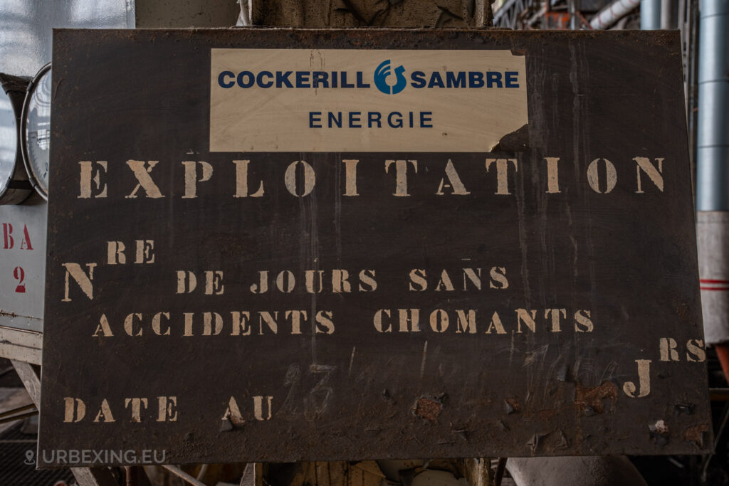 a photograph taken in at an urbex location called blue power plant. the photograph shows a rusty metal sign containing the logo of cokcerill-sambre energie with the text "explotation nre de jours sans accident shomants".