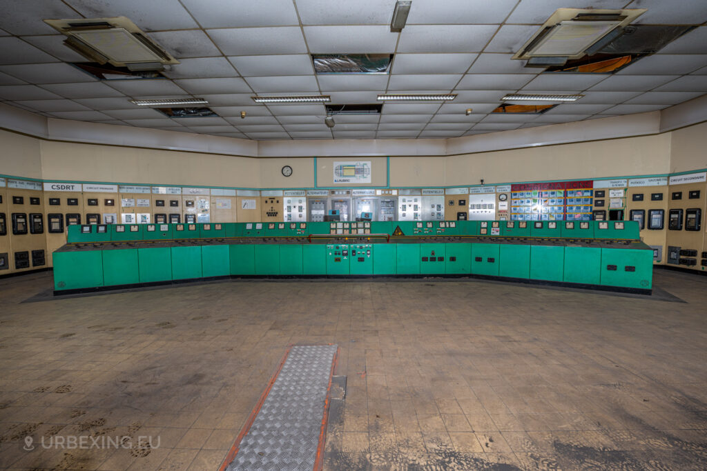 a photograph taken in at an urbex location called blue power plant. the photograph shows the control room overview, panels with meters and buttons can be seen in the walls and a large blue control panel is spreading across the room.