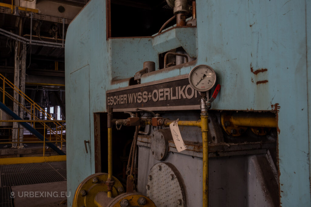 a photograph taken in at an urbex location called blue power plant. the photograph shows the logo of escher wyss and oerlikon on a blue steam turbine. there is also a pressure meter with 0, 5, 10 and 15 bar.
