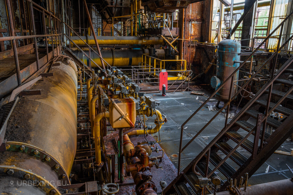 a photograph taken in at an urbex location called blue power plant. the photograph shows many gas pipes colored in yellow. a large pipe can be seen that was used to transport gas from the creation of cokes.