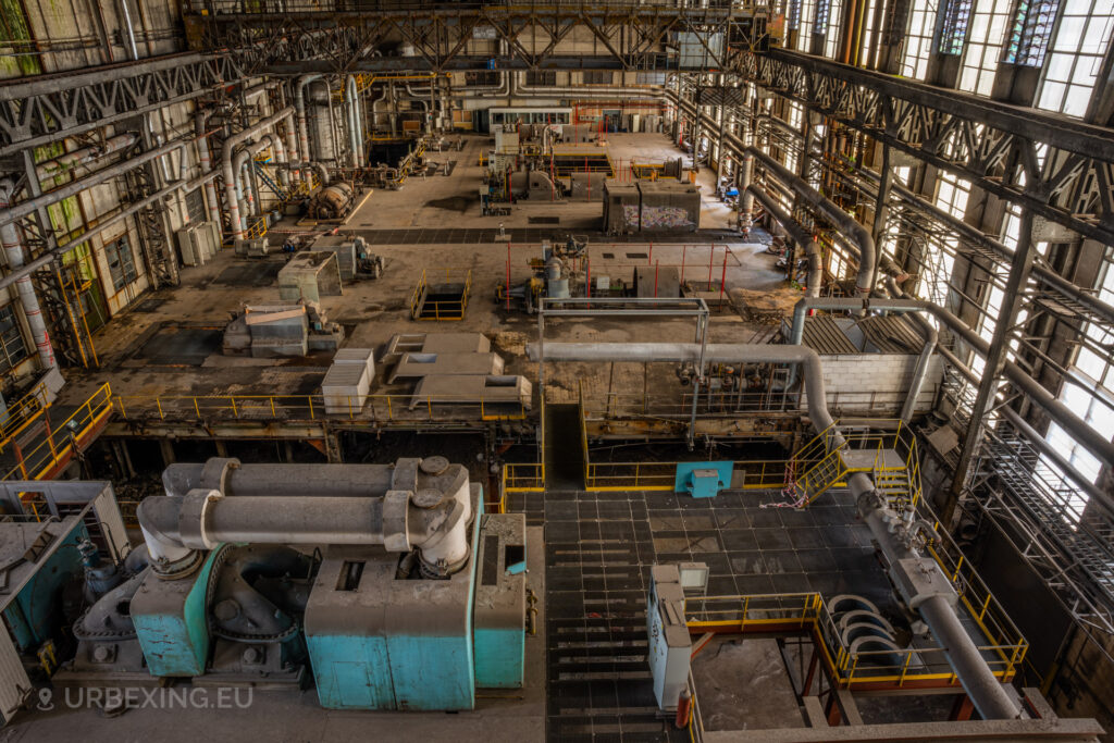 a photograph taken in at an urbex location called blue power plant. the photograph shows an overview from the top of the power plant. many turbines can be seen and a crane can be seen hanging from the ceiling.