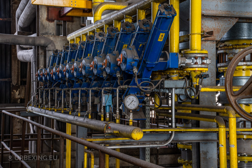 a photograph taken in at an urbex location called blue power plant. the photograph shows many hydraulic gas valves colored in yellow and blue. there is also a small pressure meter.