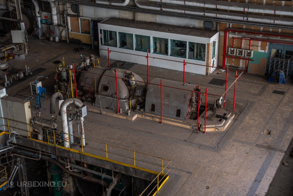 a photograph taken in at an urbex location called blue power plant. the photograph shows a small steam turbine from above with a red alternator. the railings are colored in red.