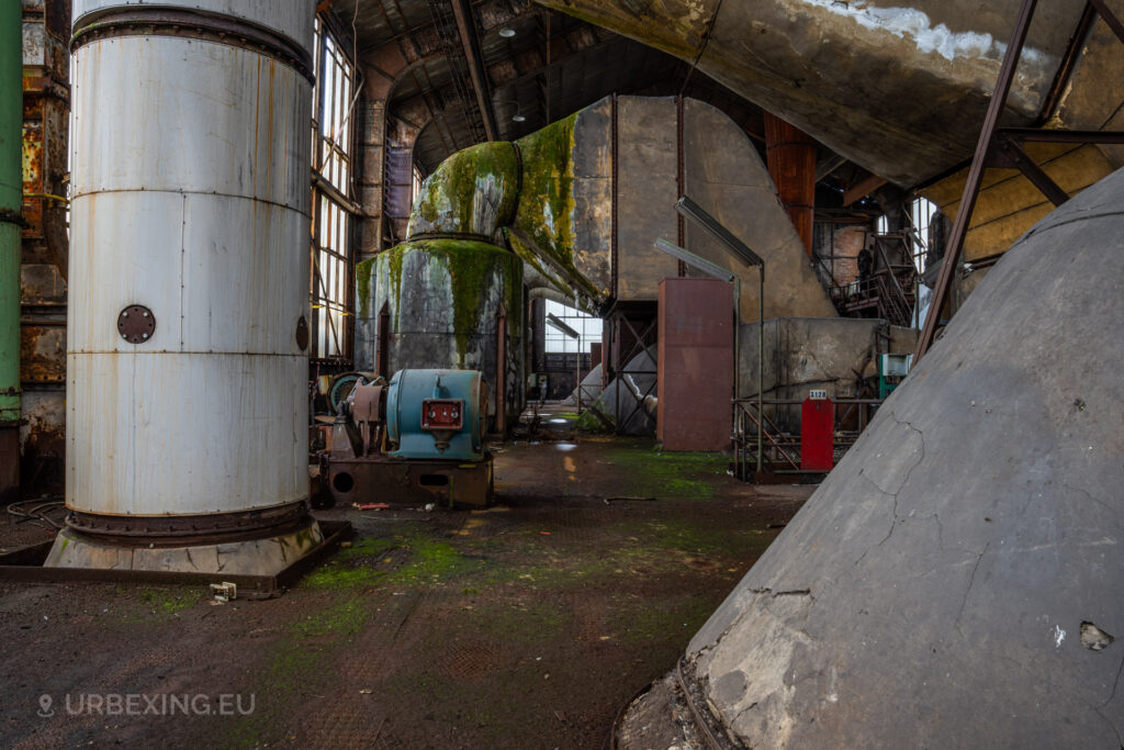 a photograph taken in at an urbex location called blue power plant. the photograph shows the top of a boiler in the boiler house. the denox installation can be seen existing of multiple pipes and a large chimney. the installation is decaying, moss and rust can be seen everywhere
