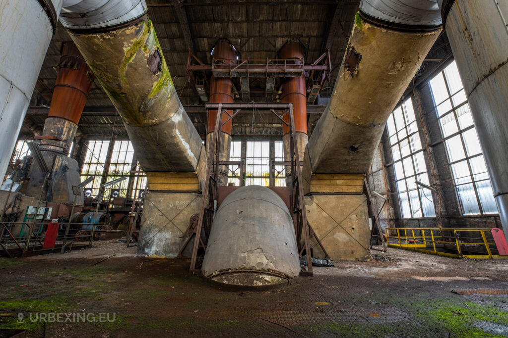 a photograph taken in at an urbex location called blue power plant. the photograph shows the top of a boiler in the boiler house. the denox installation can be seen existing of multiple pipes and a large chimney. the installation is decaying, moss and rust can be seen on the installation.