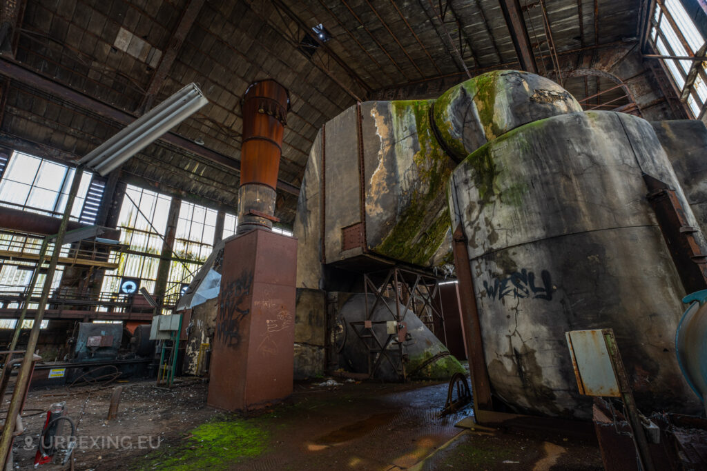 a photograph taken in at an urbex location called blue power plant. the photograph shows the top of a boiler in the boiler house. the denox installation can be seen existing of multiple pipes and a large chimney. the installation is slowly taken over by decay.