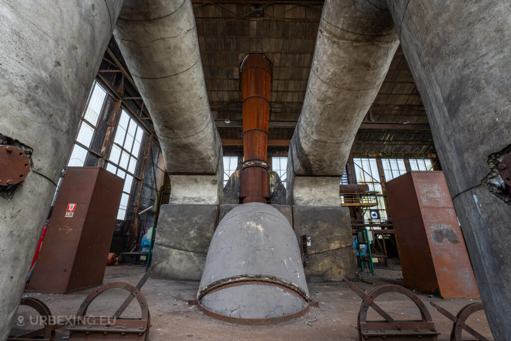 a photograph taken in at an urbex location called blue power plant. the photograph shows the top of a boiler in the boiler house. the denox installation can be seen existing of multiple pipes and a large chimney. the installation is very well preserved with little decay.
