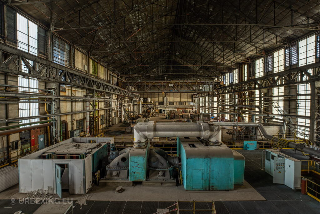 a photograph taken in at an urbex location called blue power plant. the photograph shows an overview of the turbine hall in the former power plant, a blue steam turbine can be seen along with smaller turbines in the back.