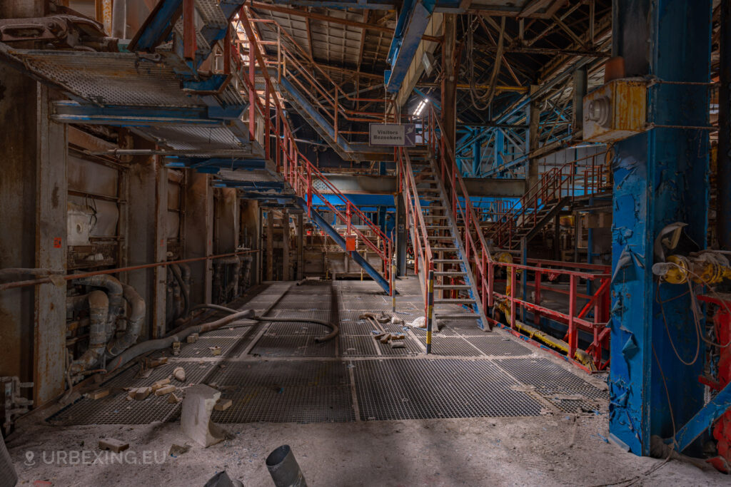 a photograph taken inside an abandoned glass factory called "de glasfabriek schiedam". The photograph shows ovens or furnaces, stairs an rails all colored in red and blue