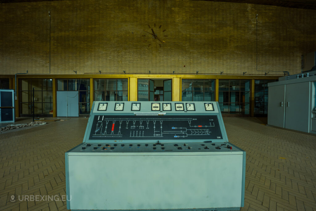 A large, old control panel with various dials and switches is positioned in the center of an abandoned radio transmitting facility. The control panel is labeled with electrical schematics and indicators. In the background, there are glass doors and windows with some broken panes, leading to what appears to be other rooms. A large, wall-mounted clock without hands is visible on the brick wall above the doors. The floor is made of light-colored tiles arranged in a herringbone pattern. The scene is dimly lit, giving a sense of abandonment and decay. The facility, known as Lost Frequencies, was discovered during urban exploring.