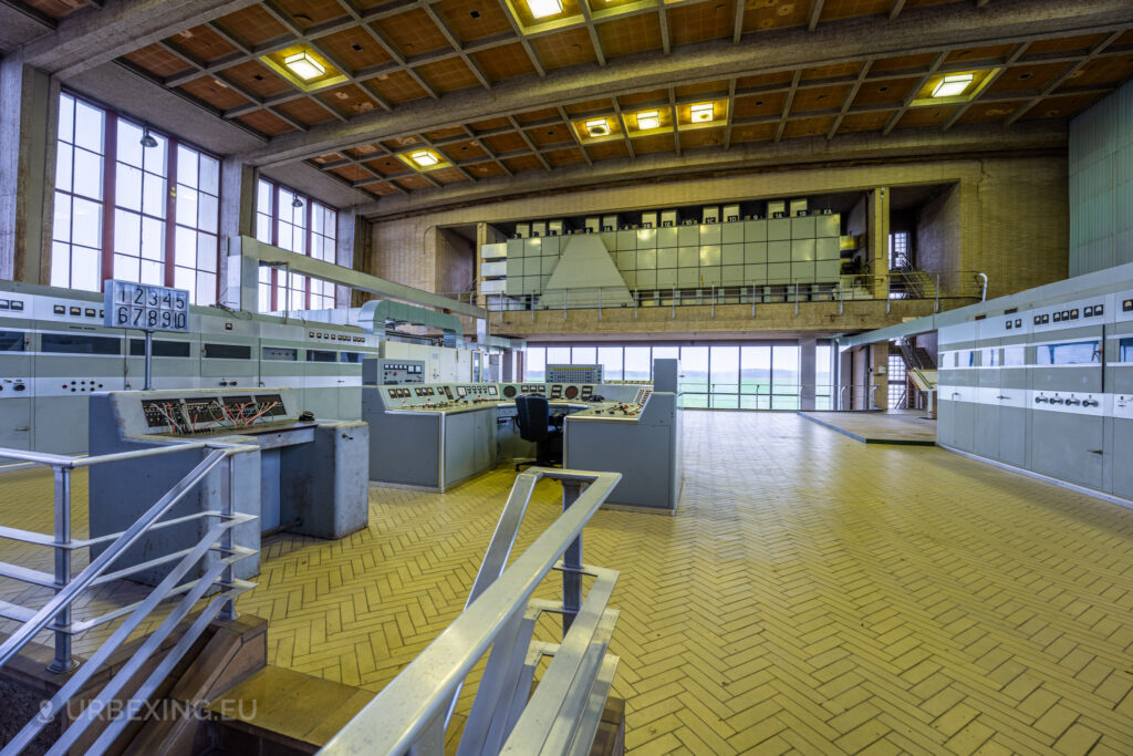 A wide shot of an abandoned control room in a radio transmitting facility, showcasing multiple control panels and machinery. The room features large windows allowing natural light to filter in, and a mezzanine level with additional equipment. The floor is tiled in a herringbone pattern, and the high ceiling has exposed beams. The overall scene conveys a sense of decay and abandonment, typical of urban exploration sites like Lost Frequencies.