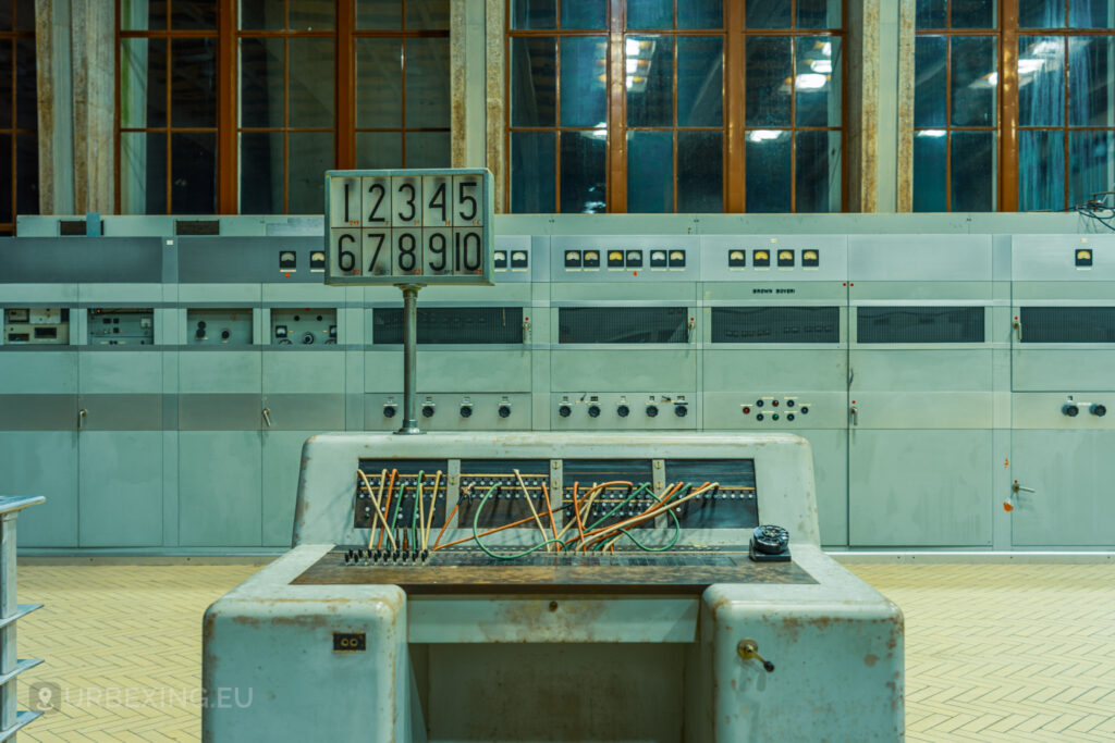 Close-up of a control panel in an abandoned radio transmitting facility, with tangled wires protruding from the front. Behind the panel, a larger wall of equipment with numerous dials and indicators is visible. The room has large windows with some broken panes, and the floor is tiled in a herringbone pattern. The scene captures the neglect and decay of the Lost Frequencies site, discovered during urban exploration.