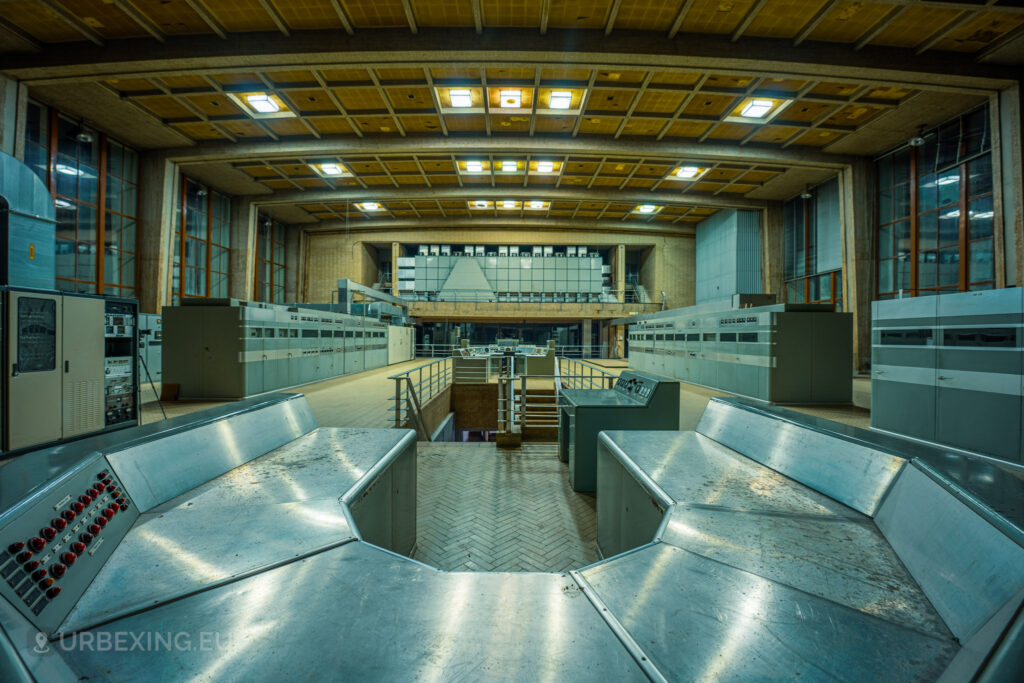 A panoramic view of an abandoned control room in a radio transmitting facility, featuring rows of control panels and equipment. The room has a high ceiling with exposed beams and large windows that let in some natural light. The central control panel is surrounded by railings and stairs, adding to the industrial feel. The floor is tiled in a herringbone pattern, and the overall atmosphere is one of desolation and abandonment, characteristic of the Lost Frequencies site explored by urban adventurers.