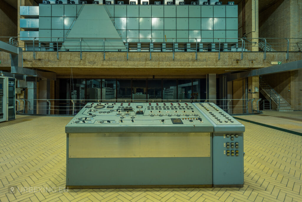 A detailed shot of a control panel in an abandoned radio transmitting facility, with numerous dials, switches, and indicators. Behind the panel, a large wall of equipment and a mezzanine level with additional machinery are visible. The room features a high ceiling with exposed beams and large windows. The floor is covered with light-colored tiles in a herringbone pattern. The scene, part of the Lost Frequencies urban exploration site, conveys a strong sense of neglect and industrial decay.