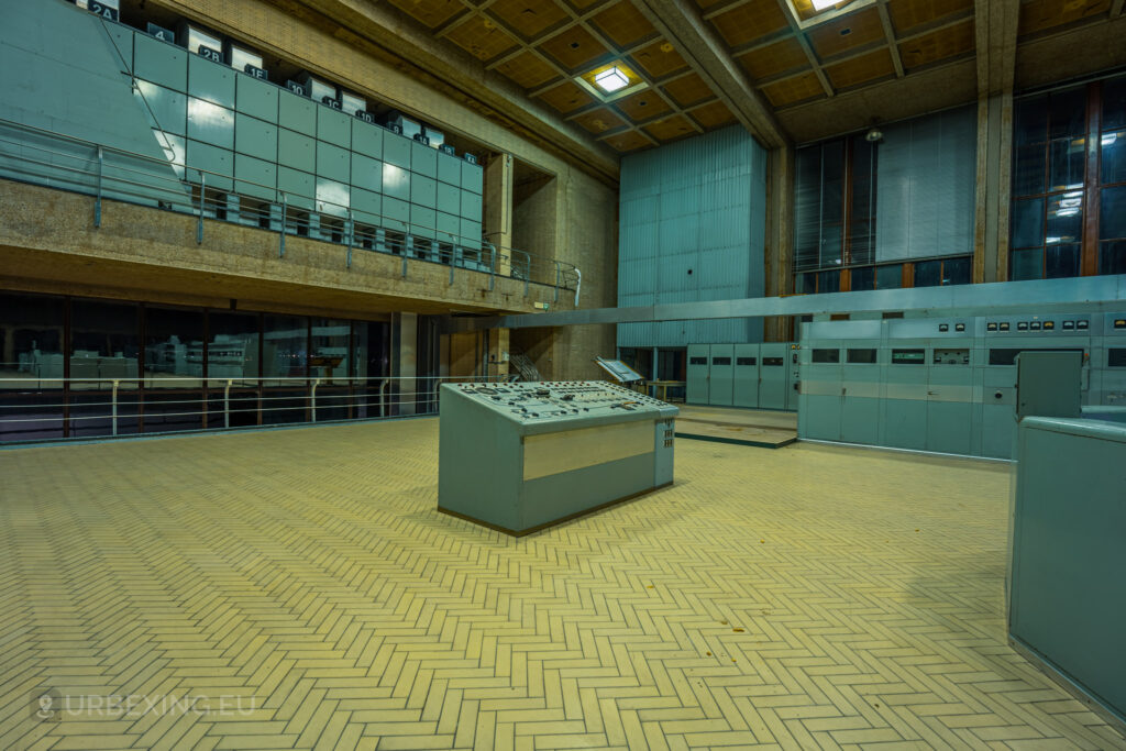 An expansive control room in an abandoned radio transmitting facility, featuring a central control panel surrounded by other machinery and equipment. The room has a high ceiling with exposed beams and large windows along one wall. Metal railings and a mezzanine level are visible, along with multiple panels labeled with numbers. The floor is covered with light-colored tiles arranged in a herringbone pattern. The space appears desolate and untouched, adding to the eerie atmosphere of the urban exploration site, known as Lost Frequencies.