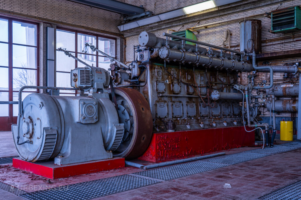 Wide shot of an abandoned diesel generator in a radio transmitting facility. The generator is rusted and shows signs of neglect, with a large flywheel and multiple pipes connected to it. The room has large windows allowing natural light to enter, and industrial equipment can be seen in the background. This image is part of the Lost Frequencies urban exploration site.