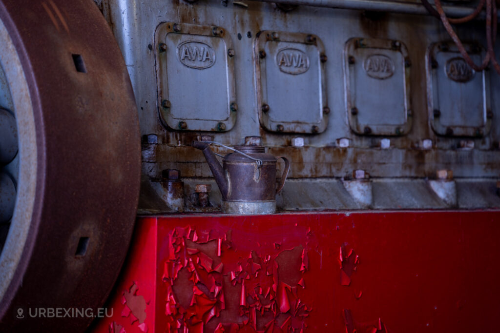 Close-up of an old diesel generator in an abandoned radio transmitting facility, focusing on the rusty engine block with the label 'AWA' visible. An old oil can is placed on the red-painted base, which has peeling paint. The scene highlights the neglect and decay of the industrial equipment at the Lost Frequencies site.