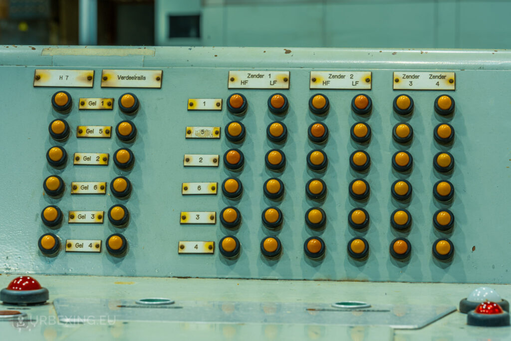 Close-up of a control panel with numerous buttons and switches in an abandoned radio transmitting facility. The panel is labeled with various indicators and functions, showing signs of wear and aging. This detailed view is part of the Lost Frequencies urban exploration site, capturing the decayed state of the equipment.