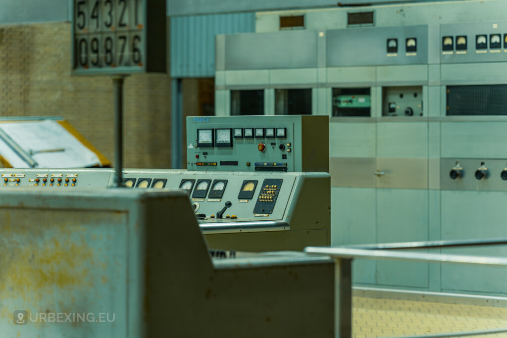 Close-up of a control panel in an abandoned radio transmitting facility, with multiple dials and switches. The panel is labeled with various indicators and buttons. Behind the panel, additional equipment and a sign with numbers can be seen. The scene is part of the Lost Frequencies urban exploration site, highlighting the abandoned and decayed state of the facility.