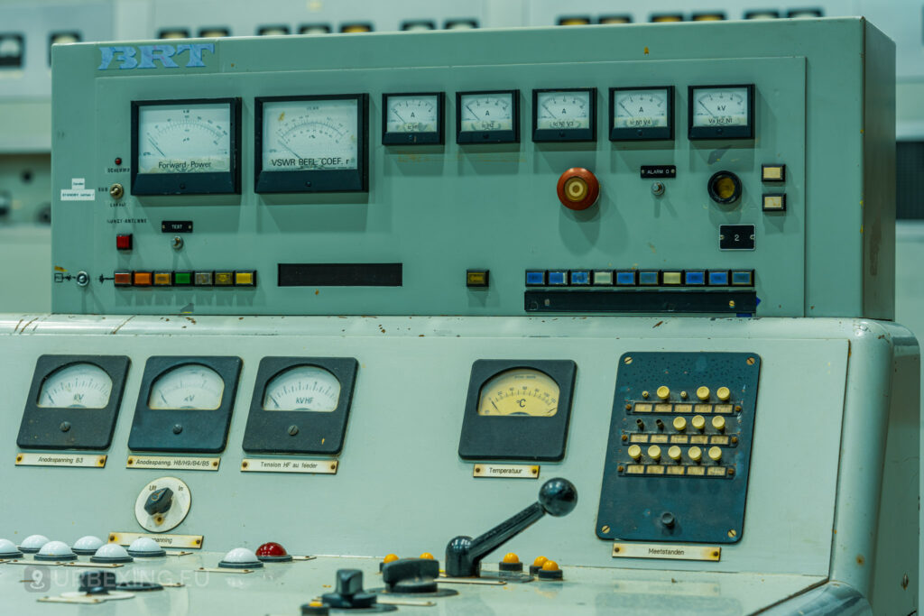 Close-up view of an old control panel in an abandoned radio transmitting facility. The panel features several analog meters, switches, and a red emergency button. Labels in multiple languages are visible, indicating different functions. The equipment is part of the Lost Frequencies site, showcasing its neglected and decayed state discovered during urban exploration