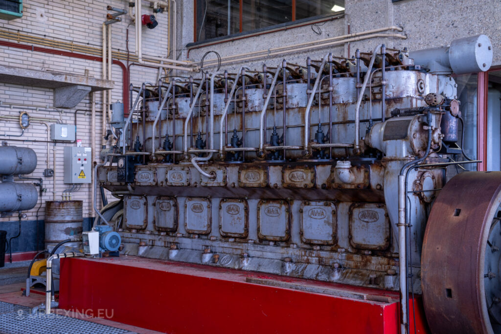 An abandoned diesel generator in a radio transmitting facility, showing signs of rust and decay. The large engine is surrounded by various pipes and cables, with a red base that has peeling paint. The background includes industrial equipment and tiled walls, emphasizing the industrial and deserted atmosphere of the Lost Frequencies site.