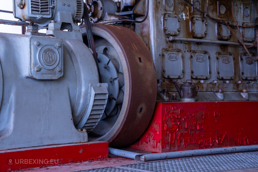 Close-up view of a diesel generator in an abandoned radio transmitting facility, focusing on the large flywheel and engine block with the label 'AWA.' The red base of the generator shows peeling paint, emphasizing the neglected state of the equipment at the Lost Frequencies site.