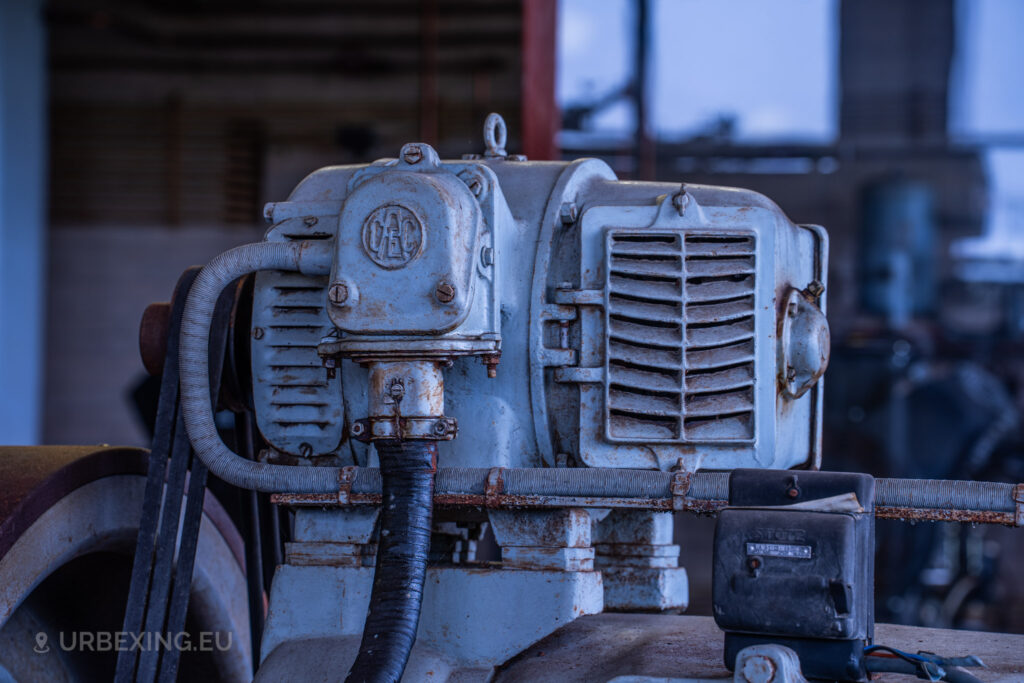 Close-up of a large diesel generator in an abandoned radio transmitting facility. The generator, showing signs of rust and decay, is situated on a red base with peeling paint. The surrounding area includes various industrial equipment, capturing the abandoned and neglected atmosphere of the Lost Frequencies site.