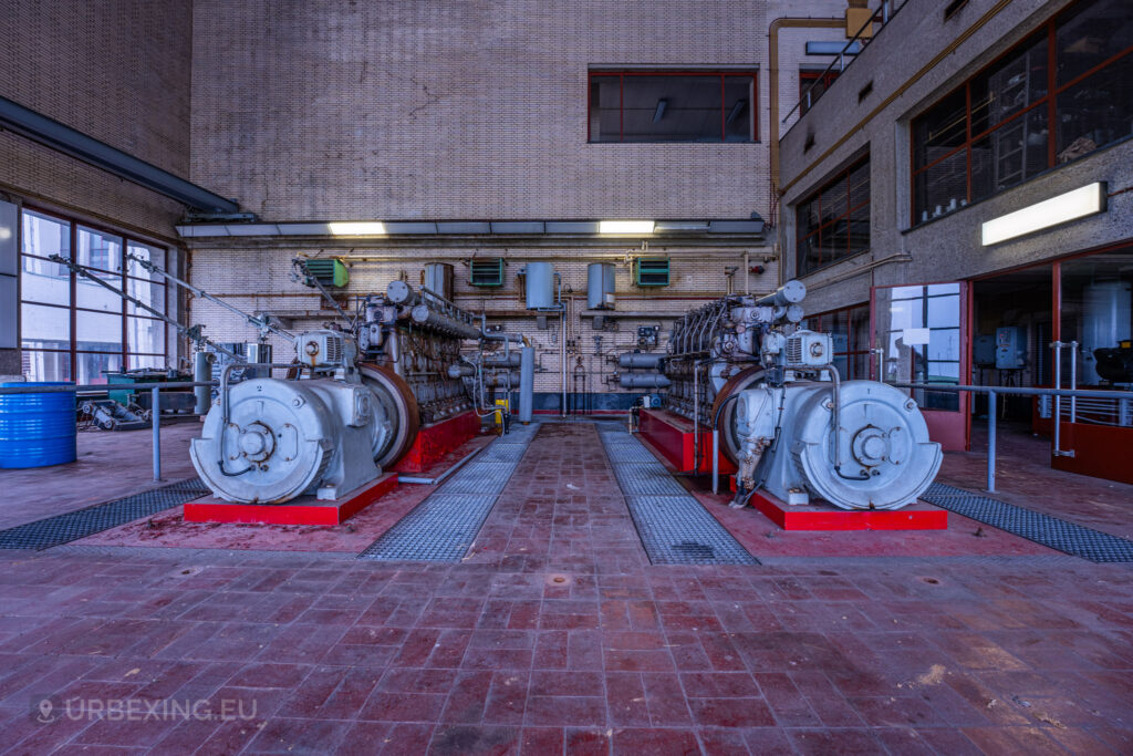 A wide shot of two large diesel generators in an abandoned radio transmitting facility. The generators are placed parallel to each other, showing signs of rust and decay. The room has a high ceiling with industrial windows and various pipes and equipment along the walls. This image is part of the Lost Frequencies urban exploration site, emphasizing the industrial and abandoned atmosphere.