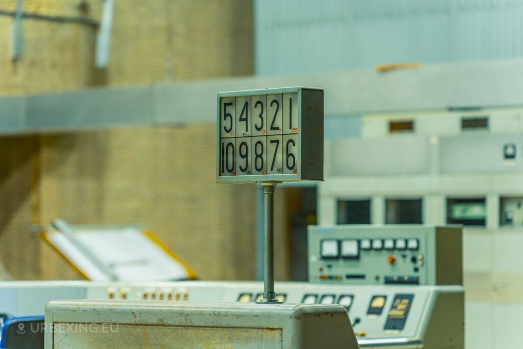 Close-up of a control panel in an abandoned radio transmitting facility, with a sign displaying numbers prominently in the foreground. The background includes additional control panels and industrial equipment. This detailed view is part of the Lost Frequencies urban exploration site, highlighting the neglected and decayed state of the equipment.