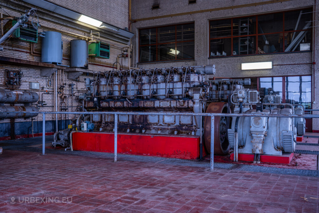 Close-up of a large diesel generator in an abandoned radio transmitting facility. The generator is rusty and shows signs of wear, with various pipes and cables connected to it. The scene emphasizes the industrial and neglected atmosphere of the Lost Frequencies site.