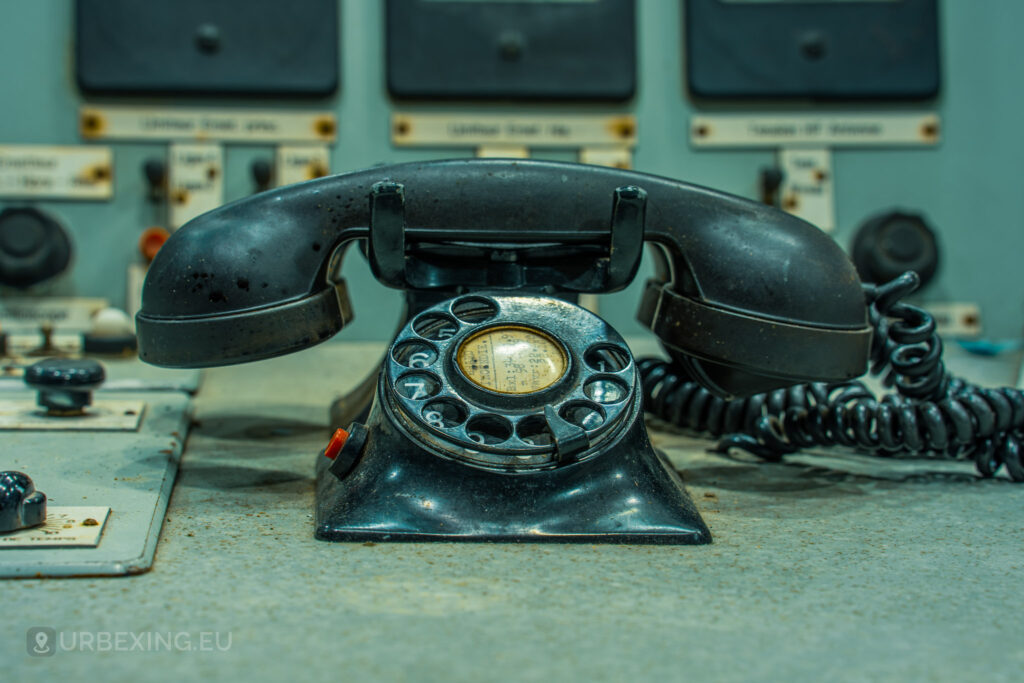 Close-up of an old rotary telephone on a control panel in an abandoned radio transmitting facility. The background includes various dials, switches, and labels. This image captures the sense of abandonment and decay at the Lost Frequencies site.