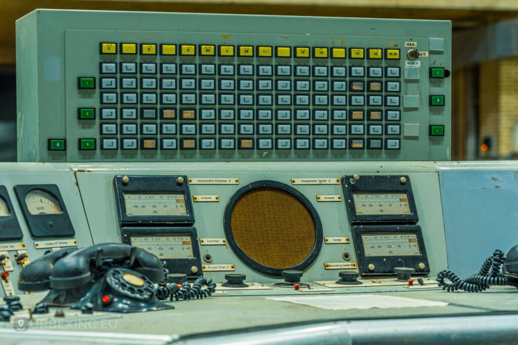 Close-up of a control panel with numerous buttons, switches, and dials in an abandoned radio transmitting facility. The panel also features a large speaker and several telephones. This detailed view highlights the industrial decay and neglected state of the Lost Frequencies site.