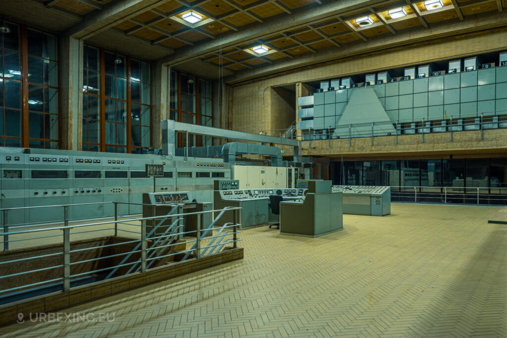 An expansive view of a control room in an abandoned radio transmitting facility. The room features a central control panel, numerous other panels with dials and switches, and a mezzanine level with additional equipment. Large windows line the walls, and the floor is tiled in a herringbone pattern. This scene is part of the Lost Frequencies urban exploration site, showcasing the abandoned and decayed state of the facility.