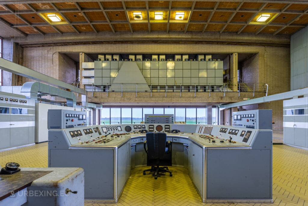 A wide shot of the control room in "Lost Frequencies," an abandoned radio transmitting facility. The image showcases a large console desk with multiple control panels, gauges, and an empty chair in the center. The architectural structure of the room, including the high ceiling and expansive windows, adds to the atmosphere of urban exploration.