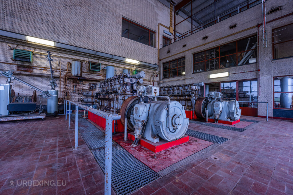 An expansive view of an industrial room in an abandoned radio transmitting facility, featuring large diesel generators and various machinery. The floor is covered with red tiles, and the high ceiling has large windows. The scene highlights the neglected and decayed state of the equipment at the Lost Frequencies site.