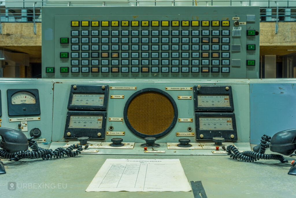 A detailed view of the control panels in the main control room of "Lost Frequencies." The image highlights various dials, switches, meters, and a central speaker. A list of handwritten notes lies on the desk, adding a human touch to the abandoned setting. Urban exploration reveals the intricate details of past operations.